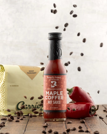 Bottle of Maple Coffee hot sauce with coffee beans and a pepper on a wooden surface.