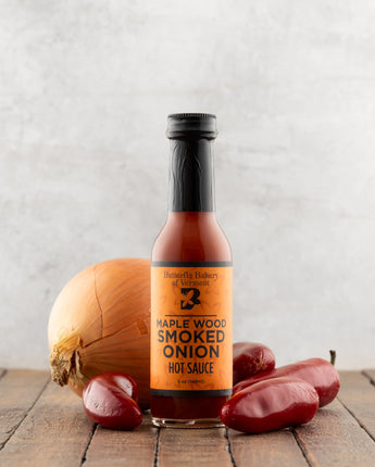 Bottle of Maple Wood Smoked Onion Hot Sauce with an onion and peppers on a wooden surface.