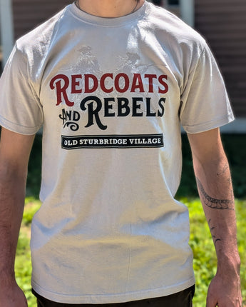 A person wearing a light beige graphic t-shirt with 'Redcoats and Rebels' and 'Old Sturbridge Village' printed on the front, photographed outdoors in front of a house and grass.