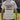 A person wearing a light beige graphic t-shirt with 'Redcoats and Rebels' and 'Old Sturbridge Village' printed on the front, photographed outdoors in front of a house and grass.